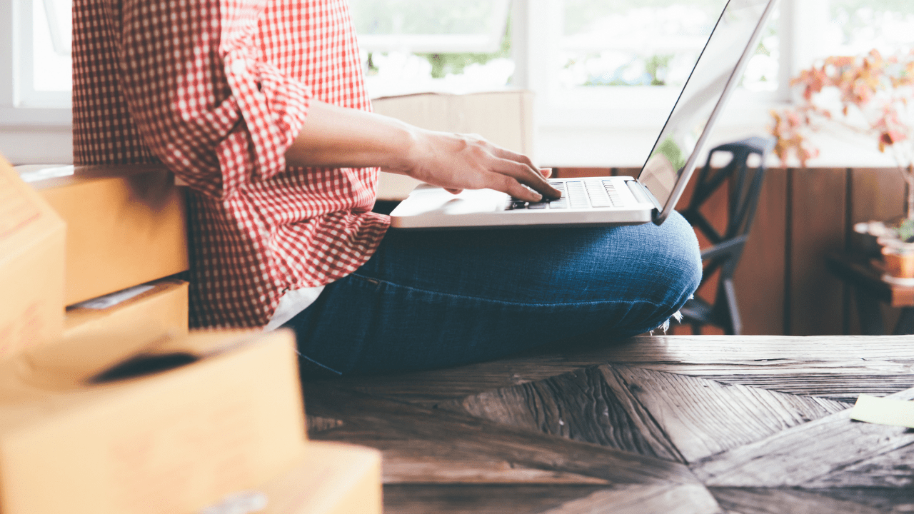 A woman in a red gingham shirt and blue jeans sits on the edge of a wooden table with a laptop in her lap while setting up online advertising for her rental property.