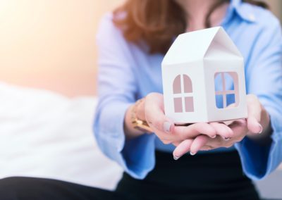 A woman in business attire wearing a blue button up shirt and black pants sits and holds a paper house in her hands.