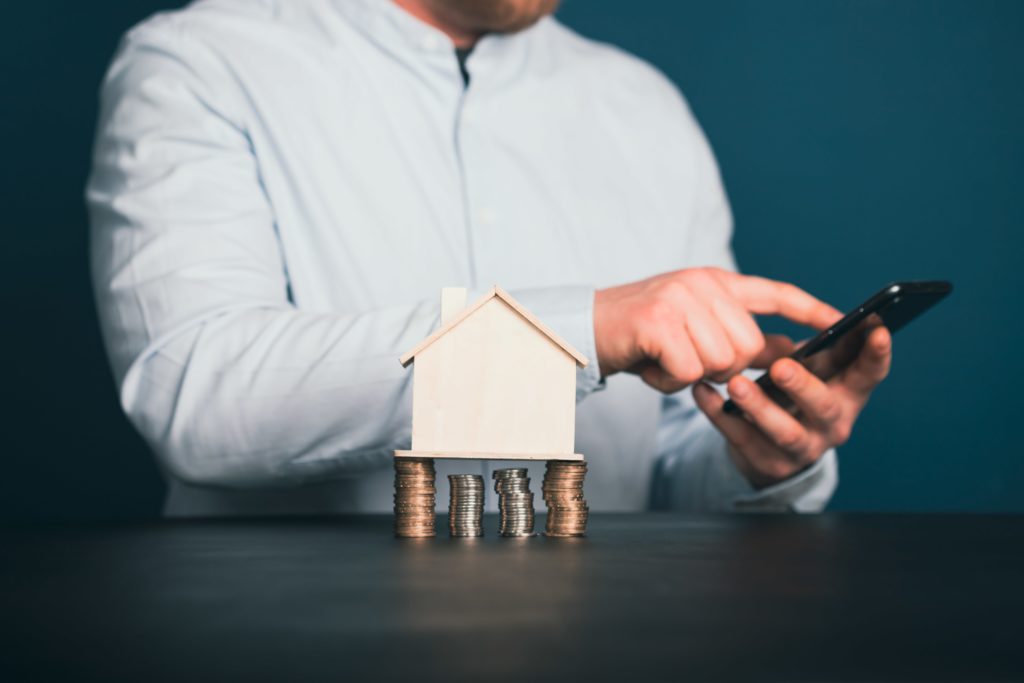 A mini fake house on top of piled up coins with a homeowner in the background looking at their phone.