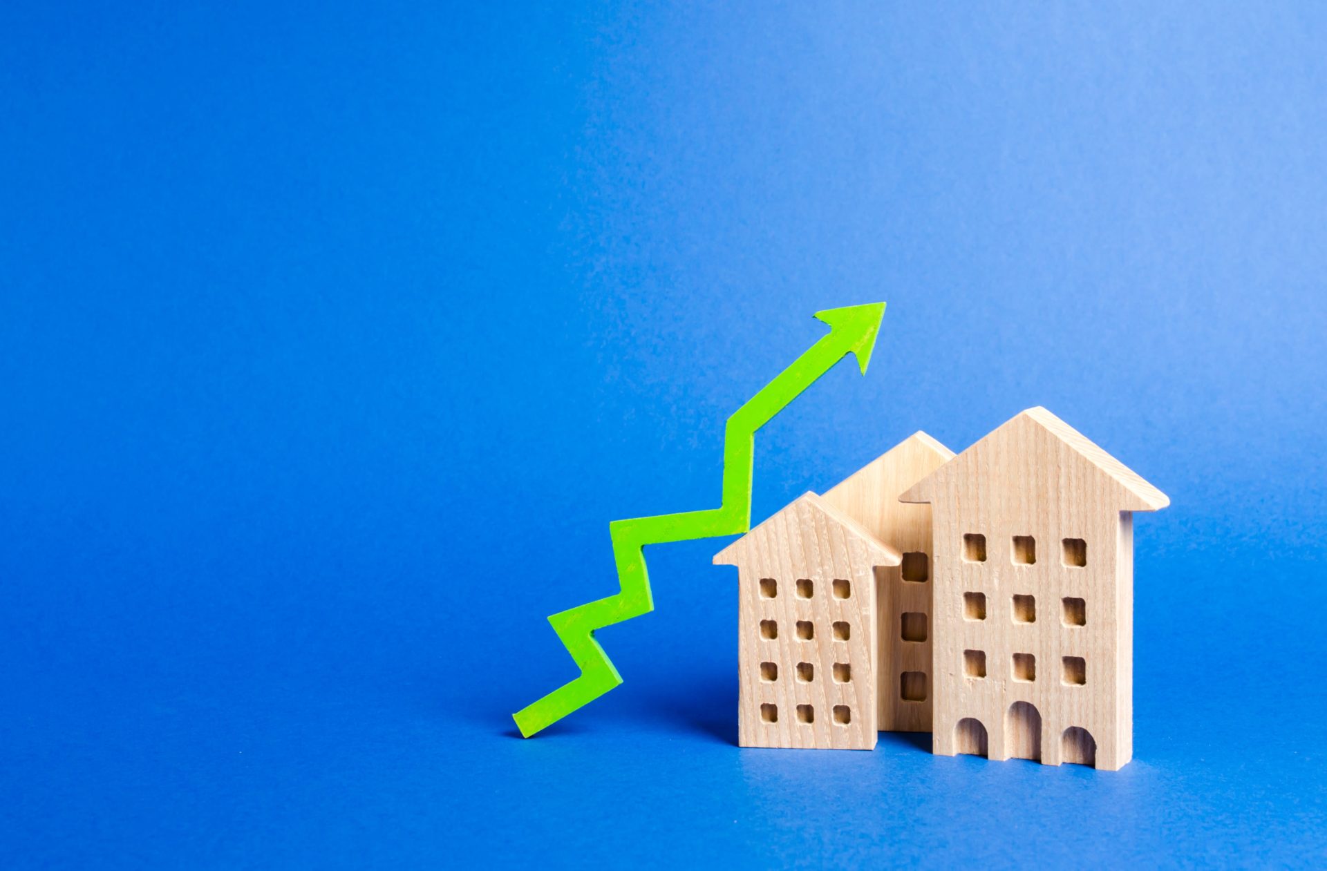 Rent Increase. An image of a wooden toy house on a blue background with a green arrow pointing up above it to signify rent increasing on a Kelowna rental.