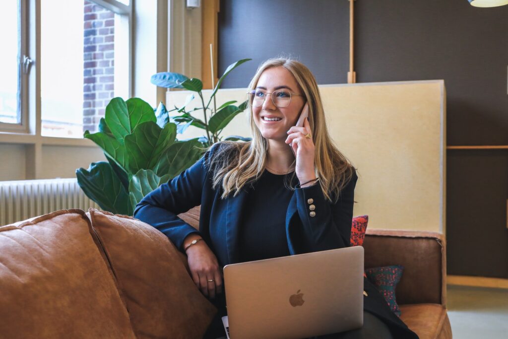 A business woman with blonde hair smiles while talking on the phone and sitting on a leather coach with a laptop on her lap.