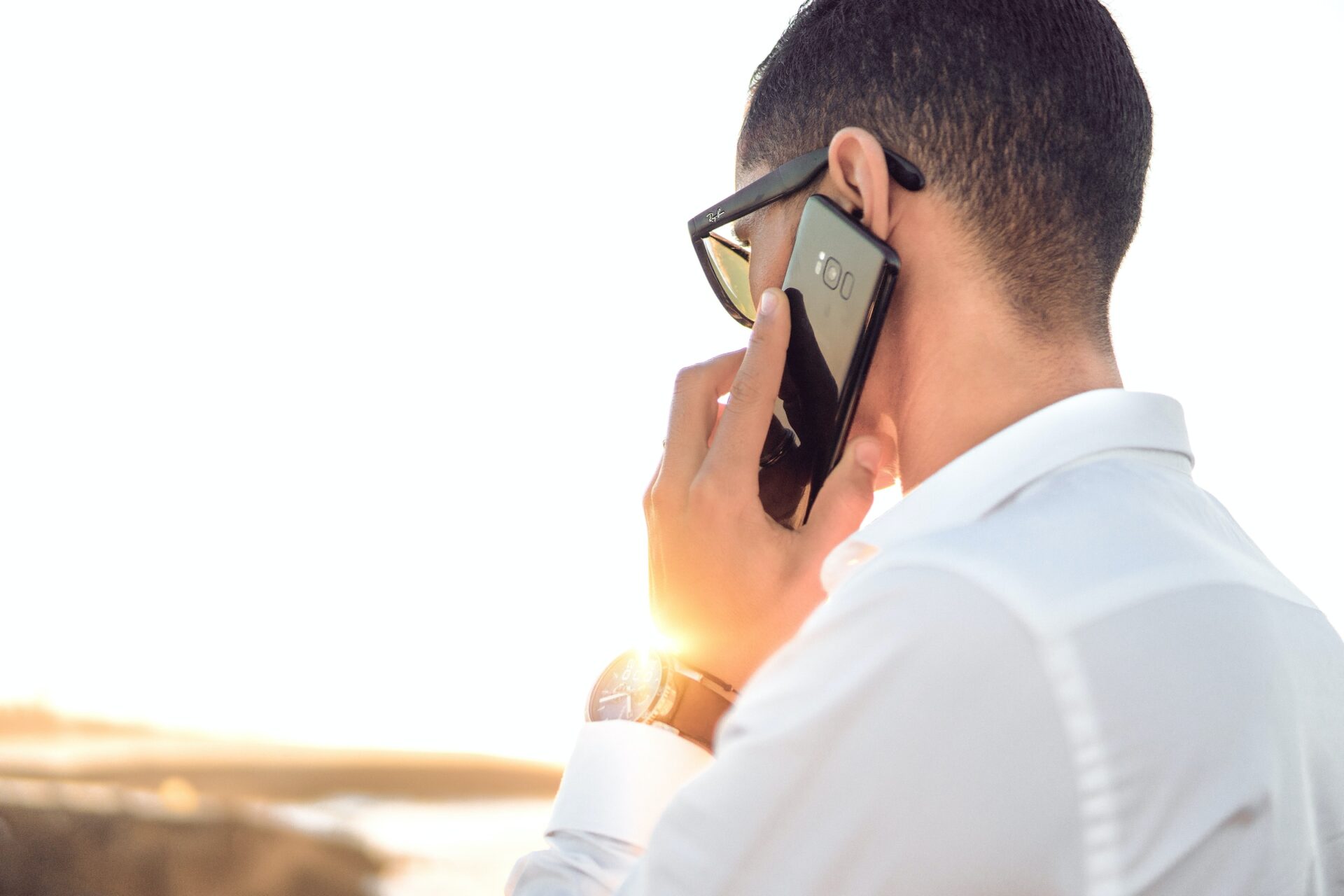 A man in a white shirt with his back turned holds a cellphone to his ear while he makes a call outside on a sunny day.