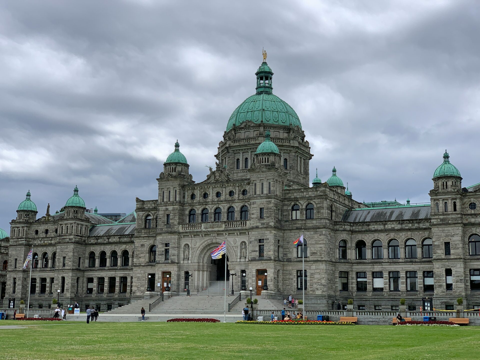 The BC Government Parliament building in Victoria BC, a large building with green roofs and the British Columbia and Canadian flags posted on a cloudy day.