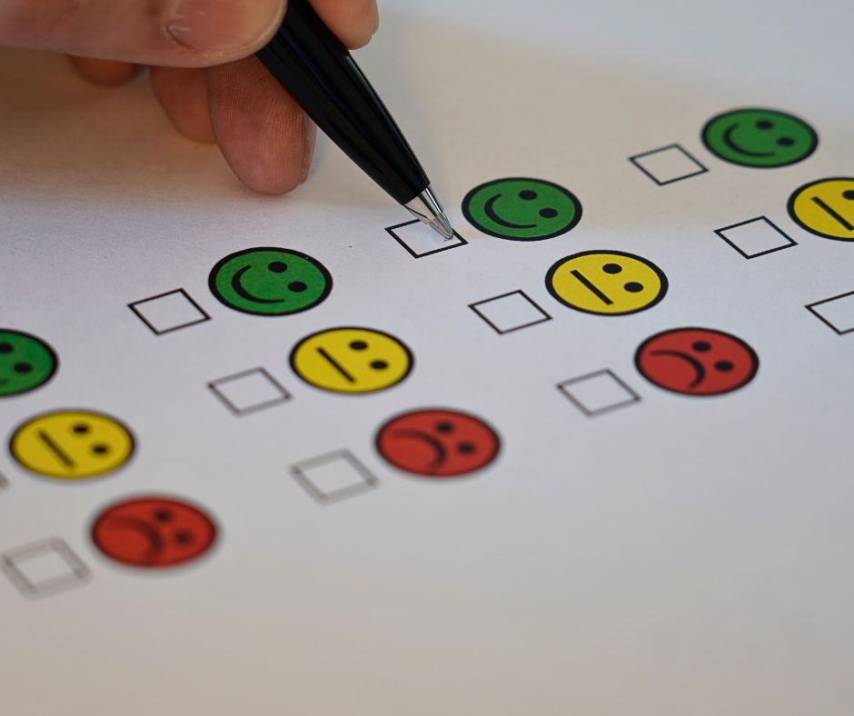 A person's hand holds and pen and fills out a feedback and review form which has three statuses to choose from, including a green happy face, a yellow neutral face, and a red angry face.