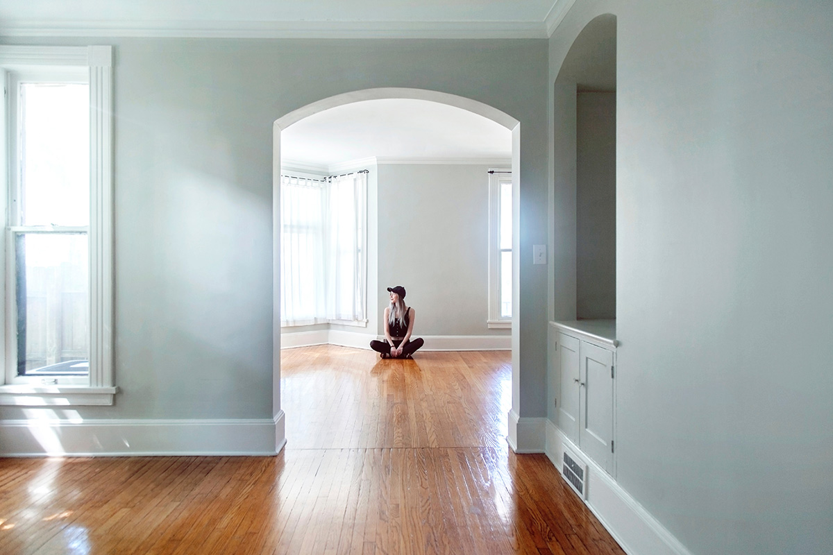 An empty, unfurnished rental with a young woman visible through an archway sitting on the floor cross legged