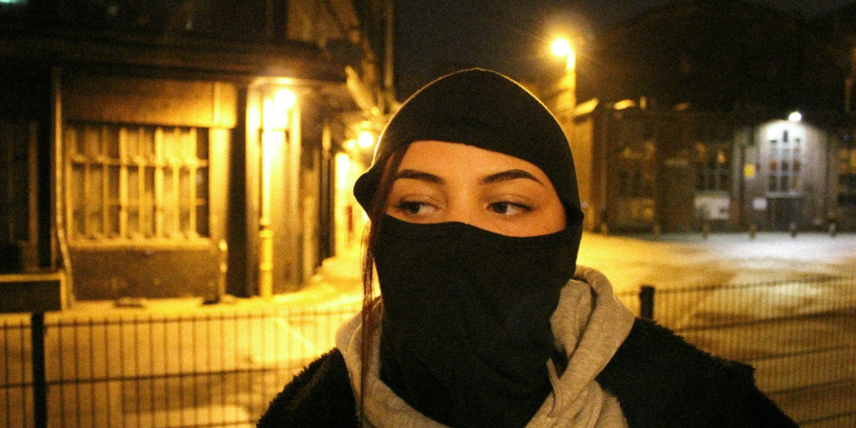 A young woman wearing a balaclava on the street at night. 
