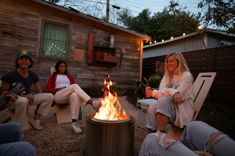 Young adults sitting around a gas fire pit in a backyard.